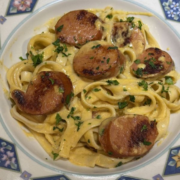 Slices of andouille sausage on fettucine alfredo, garnished with fresh parsley in a decorative bowl.