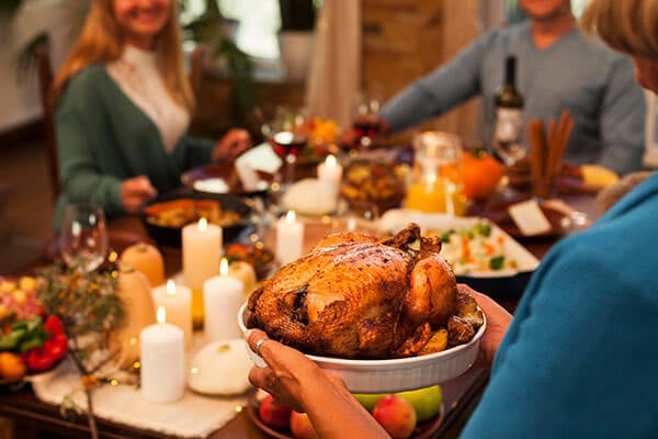 A Thanksgiving table with candles and fall decor with people seated and a person holding a cooked whole turkey.