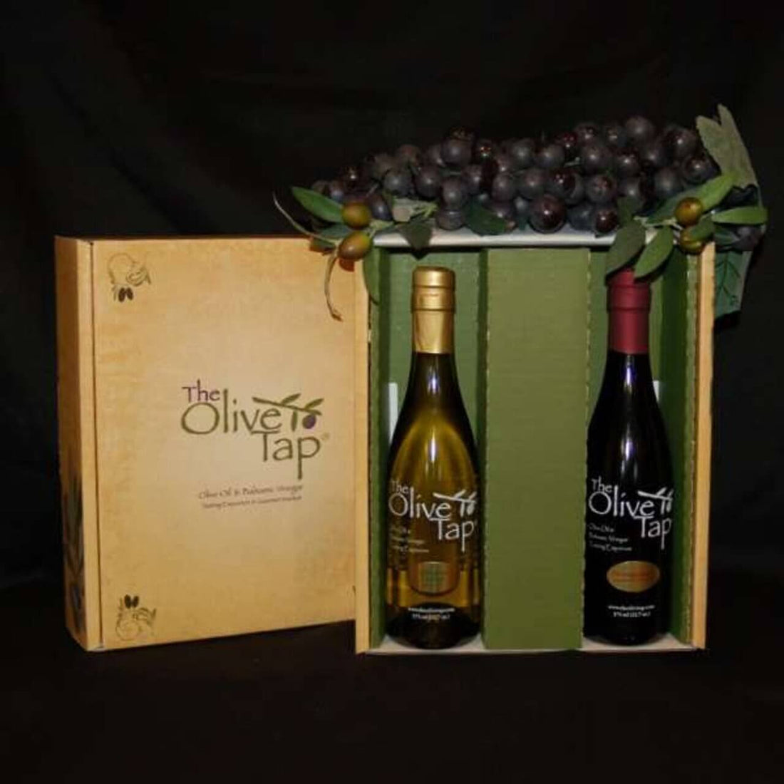 A decorative gift box from The Olive Tap with a bottle of olive oil and a bottle of vinegar inside, topped with decorative purple grapes against a black background.