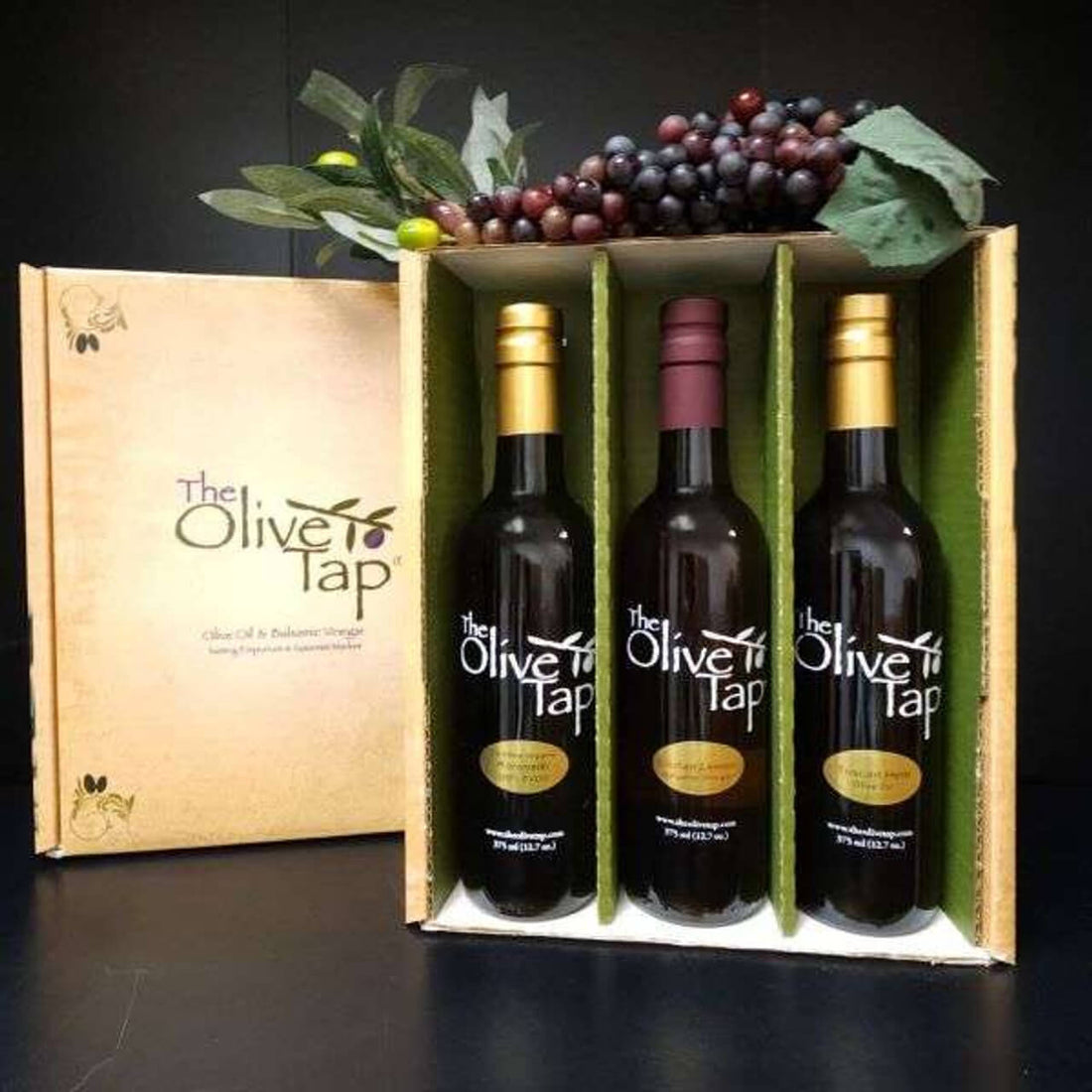 Olive Tao gift box with three bottles of olive oil and vinegar inside, separated by dividers and grapes on top against a black background and surface.