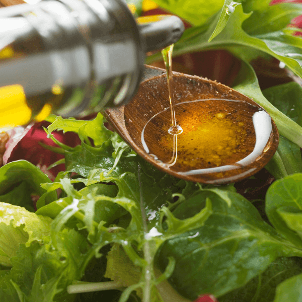 Olive oil being poured into a wooden spoon over a fresh green salad with mixed leafy greens.