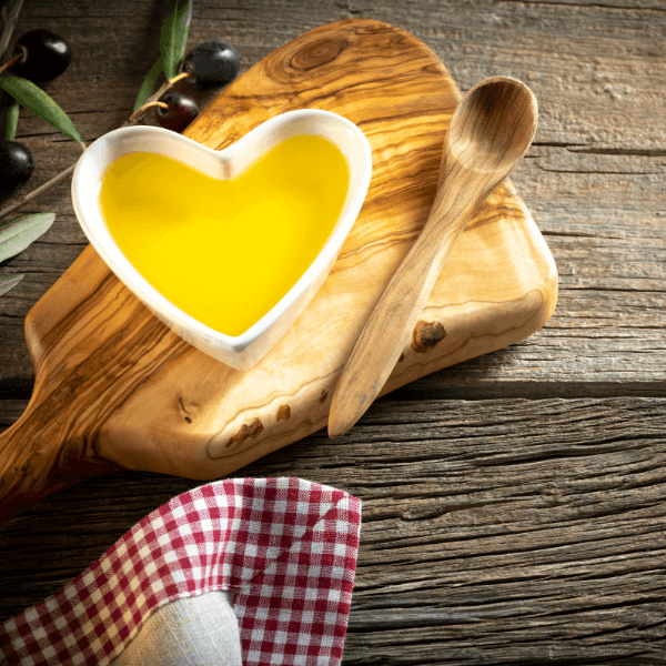 A heart-shaped bowl filled with olive oil, resting on a wooden cutting board beside a wooden spoon and olives with their branches.