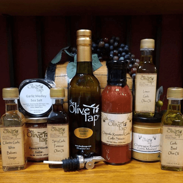 An assortment of products from The Olive Tap including olive oils, vinegars, sauces, spreads, a jar of seasoning, and a metal pour spout on a wooden surface in front of a decorative wooden barrel with purple grapes on top.