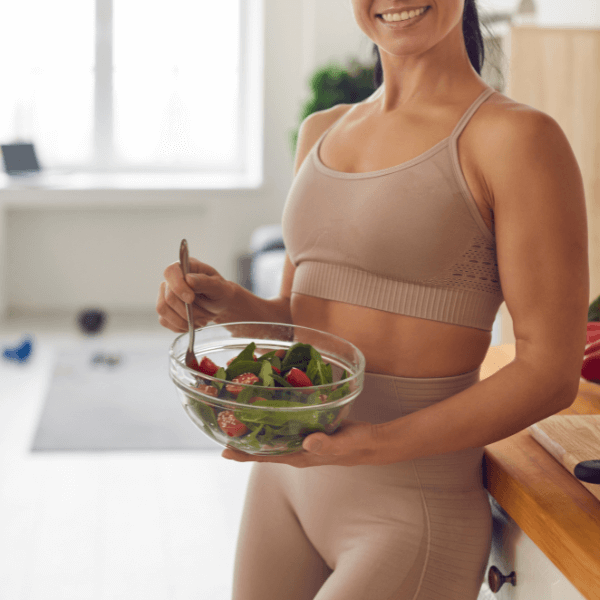 A woman in a tan-colored workout outfit leaning against a wooden countertop with a bowl of salad in one hand and a fork in the other.