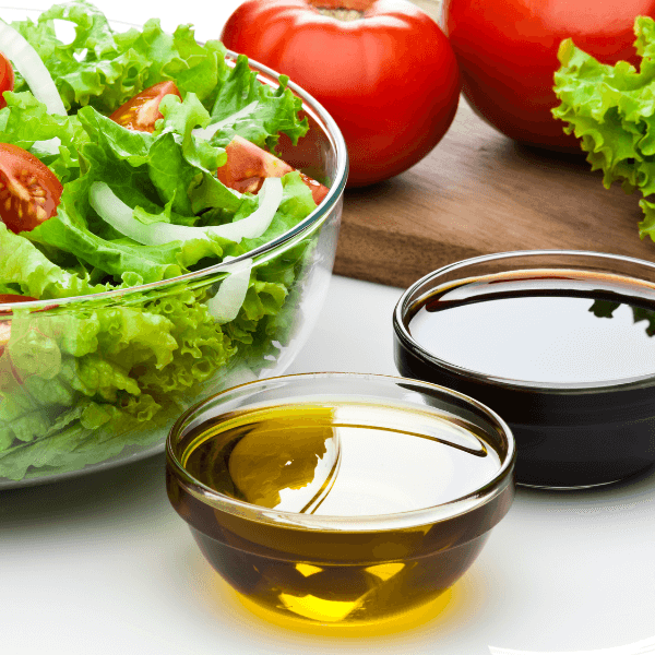 A small glass bowl of olive oil and one of balsamic vinegar next to a salad bowl with leafy greens, sliced onion, and tomatoes, and a wooden cutting board with fresh tomatoes and lettuce, all on a white surface.