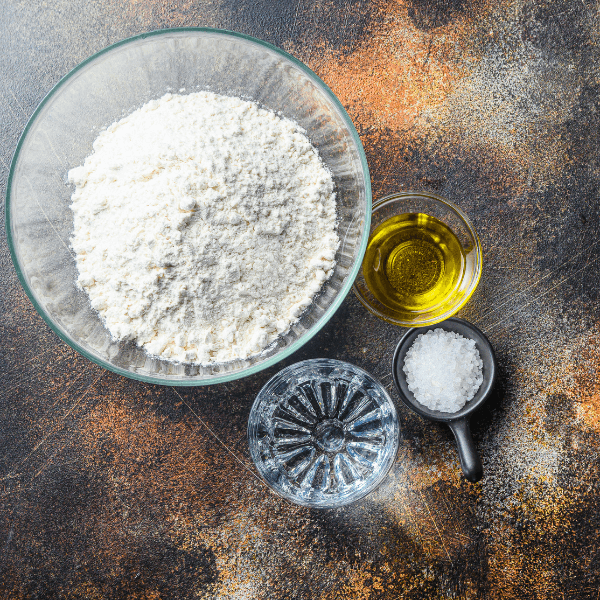 A bowl of flour with olive oil, coarse salt, and water arranged on a countertop.