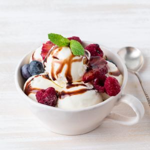 Scoops of vanilla ice cream drizzled with balsamic vinegar and topped with fresh berries, garnished with a mint leaf in a white mug with a spoon on a white surface.