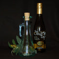 A bottle of olive oil from The Olive Tap next to a decorative empty olive oil bottle with a cork in front of olives and leaves on a dark background.