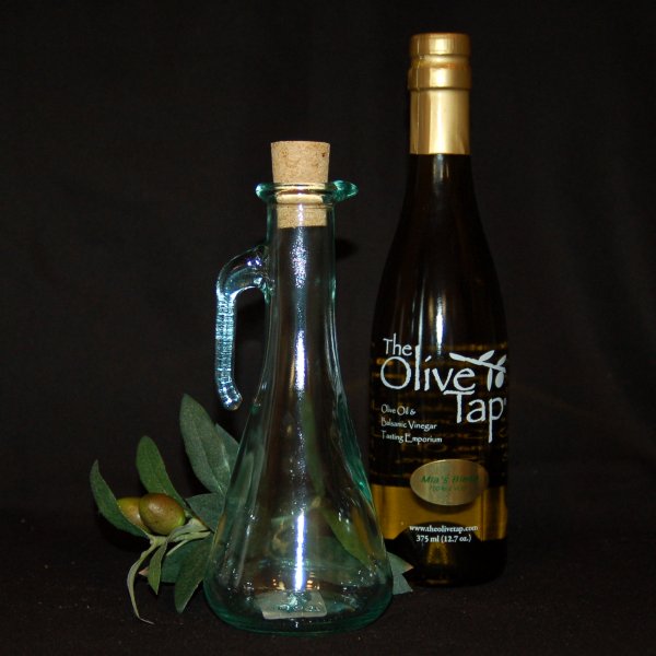 A bottle of olive oil from The Olive Tap next to a decorative empty olive oil bottle with a cork in front of olives and leaves on a dark background.