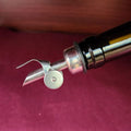 Silver olive oil pour spout in the opening of a glass bottle in front of a maroon background.
