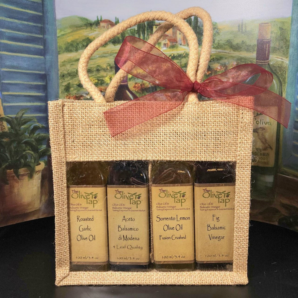 You Pick 4 100ml Sampler Set from The Olive Tap: Two bottles of olive oil and two bottles of vinegar in a decorative canvas bag with a bow with an Italian countryside mural in the background.