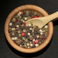 5 Peppercorn Blend In Jar from The Olive Tap: A blend of red, brown, green, black, and white peppercorns in a wooden bowl with a wooden spoon in it against a black background.