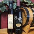 Two bottles of Apple Cider Vinegar from The Olive Tap on a wooden surface in front of a wooden barrel in front of leaves and a red background.