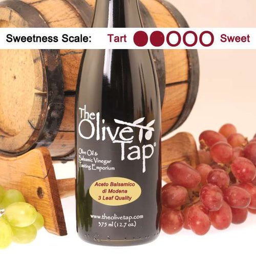 Bottle of Aceto Balsamico di Modena, 3 Leaf Quality from The Olive Tap surrounded by grapes in front of a wooden barrel, and a sweetness scale indicating a two.