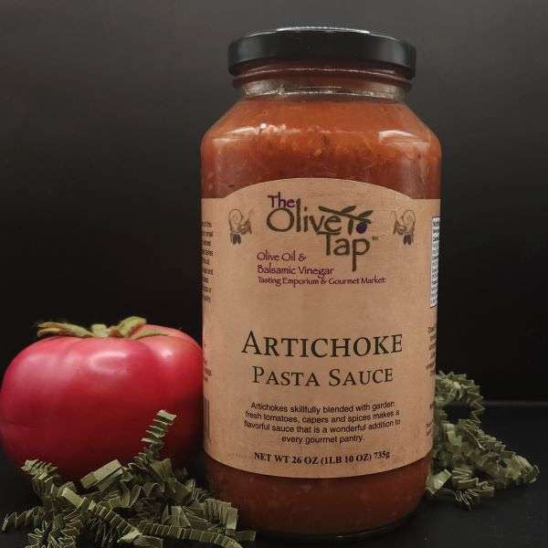 Artichoke Pasta Sauce from The Olive Tap with a fresh tomato on the left and surrounded by green decorative crinkle paper on a black surface.