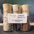 Assorted Sea Salt Trio from The Olive Tap: Three glass bottles of different colored sea salts sealed with corks and labeled on a dark surface and green background.