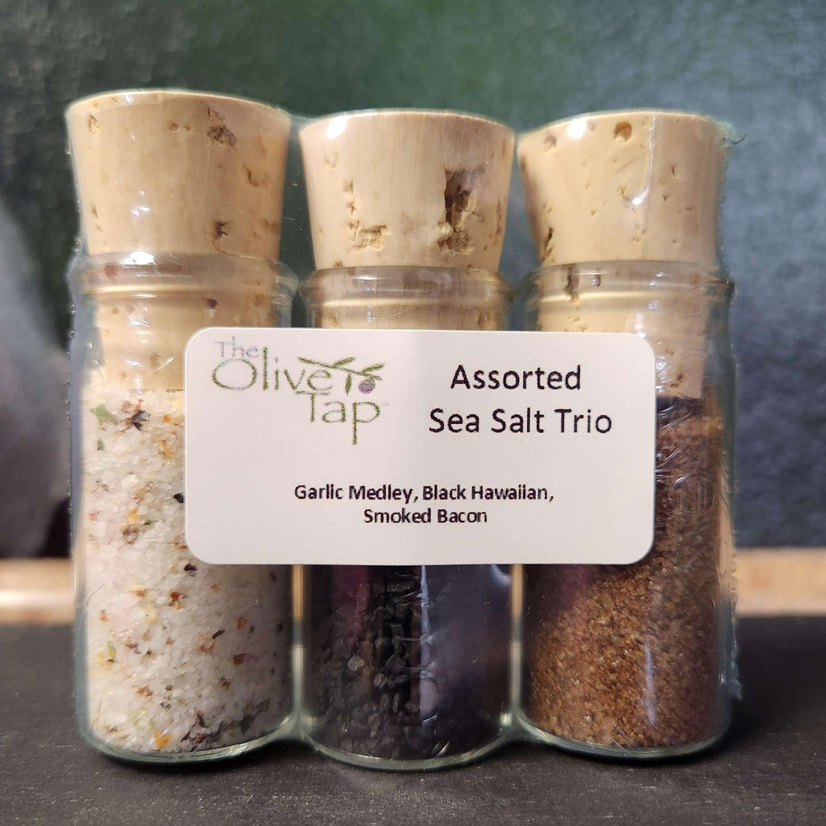 Assorted Sea Salt Trio from The Olive Tap: Three glass bottles of different colored sea salts sealed with corks and labeled on a dark surface and green background.