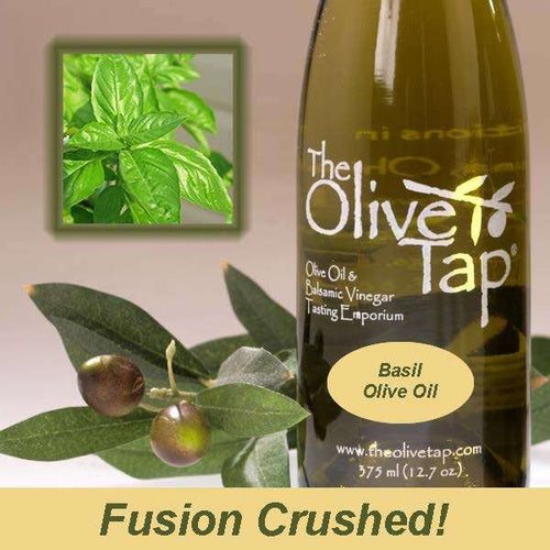 A bottle of The Olive Tap Basil Olive Oil next to olives and leaves with an inset showing a fresh basil on a light background with the words "fusion crushed!" at the bottom of the picture.