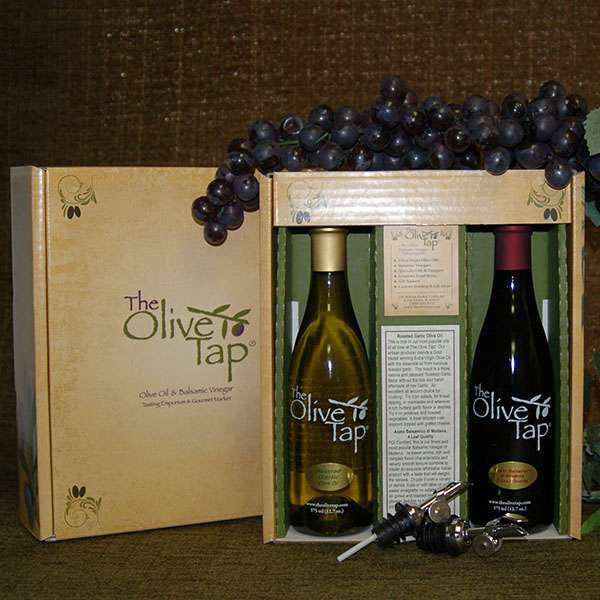 The Best Sellers Gift Box from The Olive Tap: one bottle of olive oil and one bottle of balsamic vinegar in a decorative Olive Tap box with purple grapes on top of it, divided by a piece of cardboard with a description of each product, and two metal pour spouts crossed in front of the box.