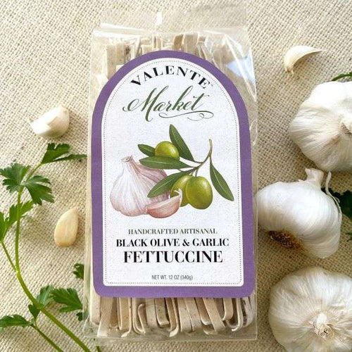 A package of Black Olive & Garlic Fettuccine from The Olive Tap on a light canvas background with fresh parsley and bulbs of garlic.