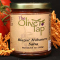 A jar of Blazin' Habanero Salsa from The Olive Tap, surrounded by orange decorative crinkle paper and tomatoes on vines behind the jar on a black surface.
