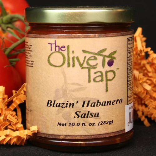 A jar of Blazin' Habanero Salsa from The Olive Tap, surrounded by orange decorative crinkle paper and tomatoes on vines behind the jar on a black surface.