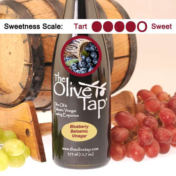 Bottle of Blueberry Balsamic Vinegar from The Olive Tap with a picture of fresh blueberries on it, surrounded by grapes in front of a wooden barrel indicating a sweetness scale of four.