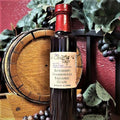 A bottle of Blueberry Chardonnay Balsamic Glaze from The Olive Tap in front of a decorative wooden barrel, purple grapes and their leaves, and a red wall in the background.