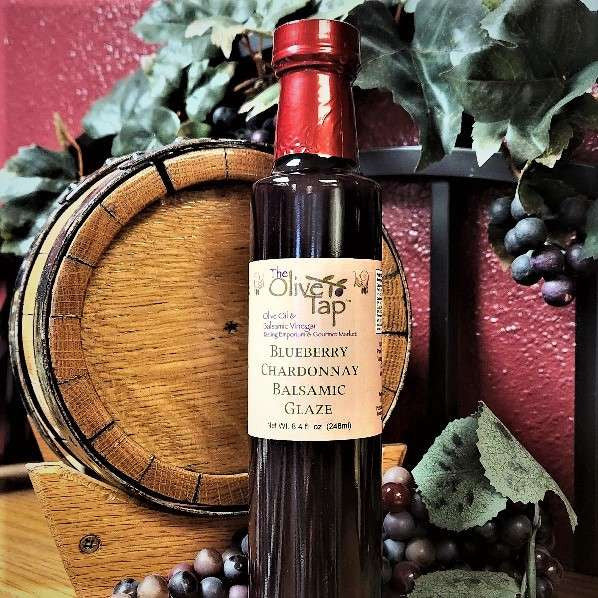 A bottle of Blueberry Chardonnay Balsamic Glaze from The Olive Tap in front of a decorative wooden barrel, purple grapes and their leaves, and a red wall in the background.