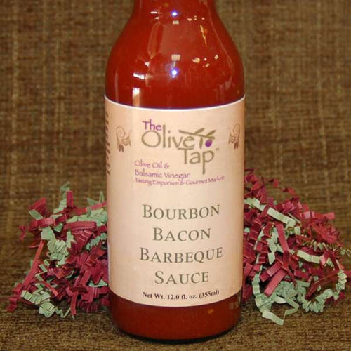 A bottle of Bourbon Bacon Barbeque Sauce from The Olive Tap in front of green and red decorative crinkle paper against a brown backdrop.