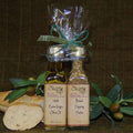Bread Dipping Pairing Set with Standard Trim from The Olive Tap: A bottle of olive oil and a bottle of bread dipping herbs tied with a bow next to bread and olives and leaves on a brown background.