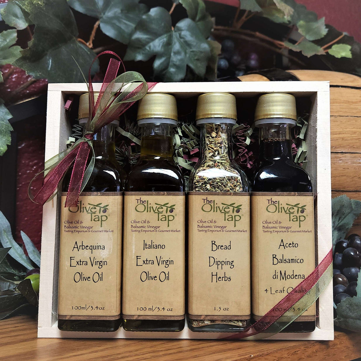 The Bread Dipping Sampler from The Olive Tap: Two bottles of olive oil, one bottle of bread dipping herbs, and one bottle of balsamic vinegar inside of a wooden box with a bow and grapes, leaves, and a wooden barrel in the background.