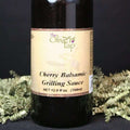 A bottle of Cherry Balsamic Grilling Sauce from The Olive Tap with green crinkle paper against a black background.