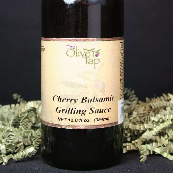 A bottle of Cherry Balsamic Grilling Sauce from The Olive Tap with green crinkle paper against a black background.