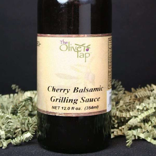 A bottle of Cherry Balsamic Grilling Sauce from The Olive Tap with green crinkle paper against a black background.