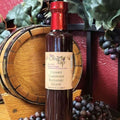 A bottle of Cherry Cabernet Balsamic Glaze from The Olive Tap in front of a decorative wooden barrel, purple grapes and their leaves, and a red wall in the background.