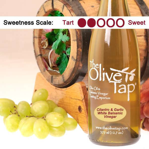 A bottle of Cilantro & Garlic White Balsamic Vinegar surrounded by grapes in front of a wooden barrel with cilantro and garlic inside, and a sweetness scale indicating a two on a light background.