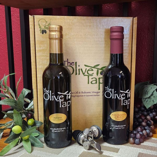 Classic Italian Gift Box from The Olive Tap: One bottle of olive oil one bottle of vinegar in front of a decorative Olive Tap box with two crossed pour spouts in the middle of the two bottles, and olive branches on the left and purple grapes on the right, all in front of a red background.