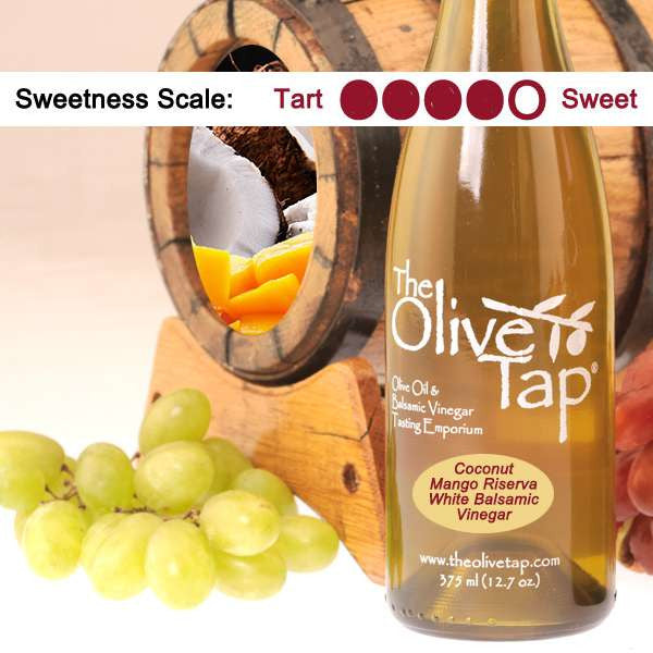 A bottle of Coconut Mango White Riserva Balsamic Vinegar from The Olive Tap surrounded by grapes in front of a wooden barrel with a picture of coconuts and mango in it, and a sweetness scale indicating a four on a light background.