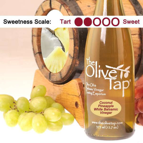 Coconut Pineapple White Balsamic Vinegar from The Olive Tap with grapes in front of a wooden barrel with a picture of a coconut and pineapple inside, and a scale indicating a sweetness of wo.