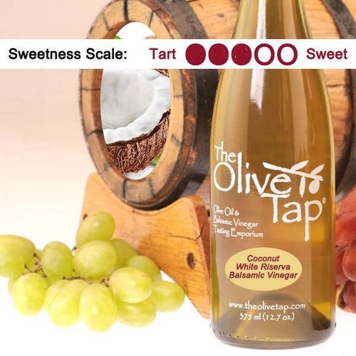 Bottle of Coconut White Riserva Balsamic Vinegar from The Olive Tap with grapes in front of a wooden barrel that has a picture of a coconut inside, indicating a sweetness scale of three.