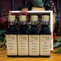 Cordial Sampler in Wood Box from The Olive Tap: Four bottles of vinegar lined up in a wooden box with a bow on a wooden surface in front of a decorative wooden barrel with olives, leaves, and grapes.