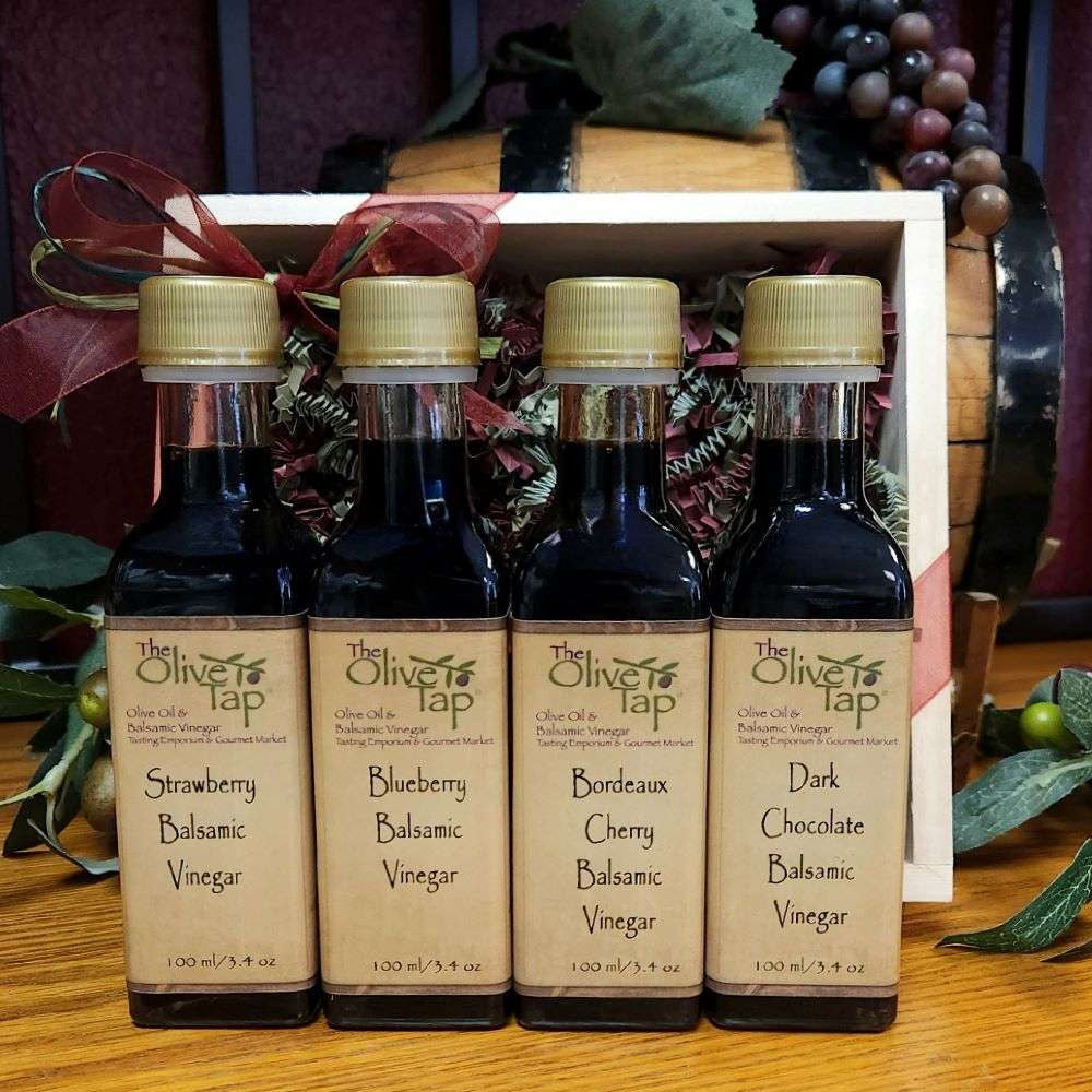 Cordial Sampler in Wood Box from The Olive Tap: Four bottles of vinegar lined up in a wooden box with a bow on a wooden surface in front of a decorative wooden barrel with olives, leaves, and grapes.