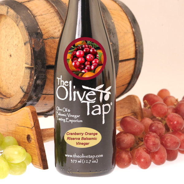 A bottle of Cranberry Orange Riserva Balsamic Vinegar from The Olive Tap with a picture of fresh cranberries and oranges, surrounded by grapes in front of a wooden barrel on a light background.