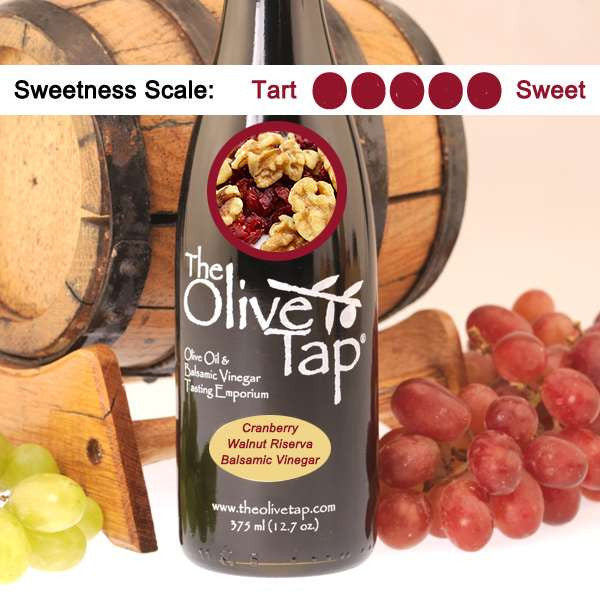 A bottle of Cranberry Walnut Riserva Balsamic Vinegar from The Olive Tap with a picture of cranberries and walnuts, surrounded by grapes in front of a wooden barrel, and a sweetness scale indicating a five on a light background.