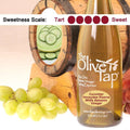 A bottle of Cucumber Honeydew Riserva White Balsamic Vinegar from The Olive Tap surrounded by grapes in front of a wooden barrel with a sliced honeydew and cucumber slices inside, and a sweetness scale indicating a five on a light background.