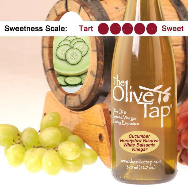 A bottle of Cucumber Honeydew Riserva White Balsamic Vinegar from The Olive Tap surrounded by grapes in front of a wooden barrel with a sliced honeydew and cucumber slices inside, and a sweetness scale indicating a five on a light background.