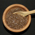 Durango Hickory Smoked Sea Salt from The Olive Tap: Brown grains of sea salt in a wooden bowl and a wooden spoon against a black background.