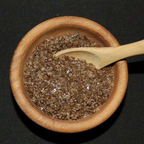 Durango Hickory Smoked Sea Salt from The Olive Tap: Brown grains of sea salt in a wooden bowl and a wooden spoon against a black background.
