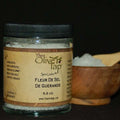 A jar of Fleur De Sel De Guerande Sea Salt from The Olive Tap with a wooden bowl and spoon full of sea salt in the background.